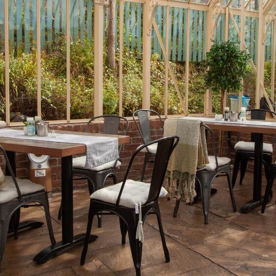 Walnut-topped tables with black bistro chairs and cream cushions inside the glasshouse.