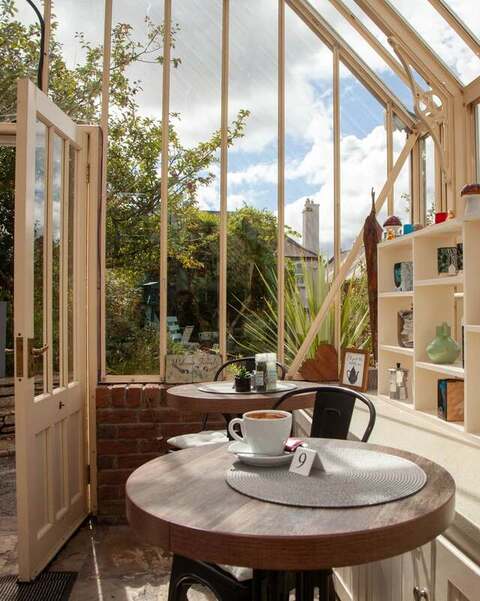 A cappuccino on table nine beside an open glasshouse door, with a shelf of cups behind.